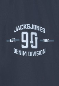 Námořnická modrá bavlněná tričko s grafikou, na které je text "JACK&JONES," "90," a "DENIM DIVISION" v bílé a světle šedé.