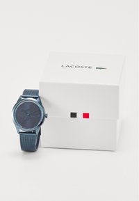 Montre Lacoste avec un bracelet en maille bleu, cadran rond bleu, mouvement à quartz et design minimaliste. Boîte blanche avec logo et accents colorés.