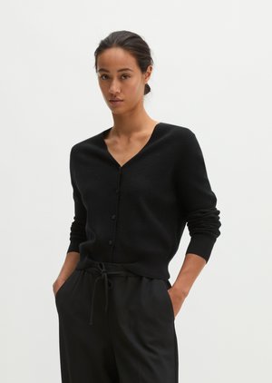 Frau trägt einen schwarzen, geknöpften Cardigan und schwarze Hose mit Kordelzug, steht mit Händen in den Taschen vor einem schlichten weißen Hintergrund.