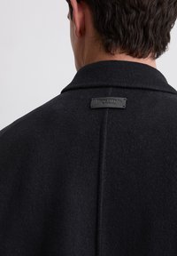 Schwarzer Wollmantel mit strukturiertem Kragen und texturiertem Finish, ausgestattet mit einem rechteckigen Stoffetikett am hinteren Nacken mit geprägtem Branding.