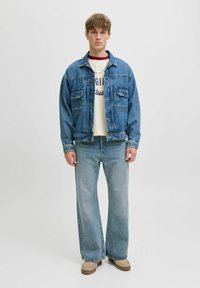 Jauns vīrietis valkā zilu denim jaku, krēmkrāsas grafisko T-kreklu, gaiši zilus platās bikses un bežīgas kurpes, stāvot pretī vienkāršai fona.