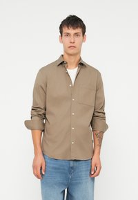 Cămașă maro cu nasturi și guler, cu un singur buzunar pe piept și mâneci răsucite, purtată peste un tricou alb, asortată cu blugi ușor albaștri.