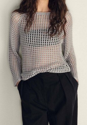 Femme portant un pull gris à manches longues en maille ajourée sur un haut noir, associé à un pantalon noir ample, mains dans les poches.