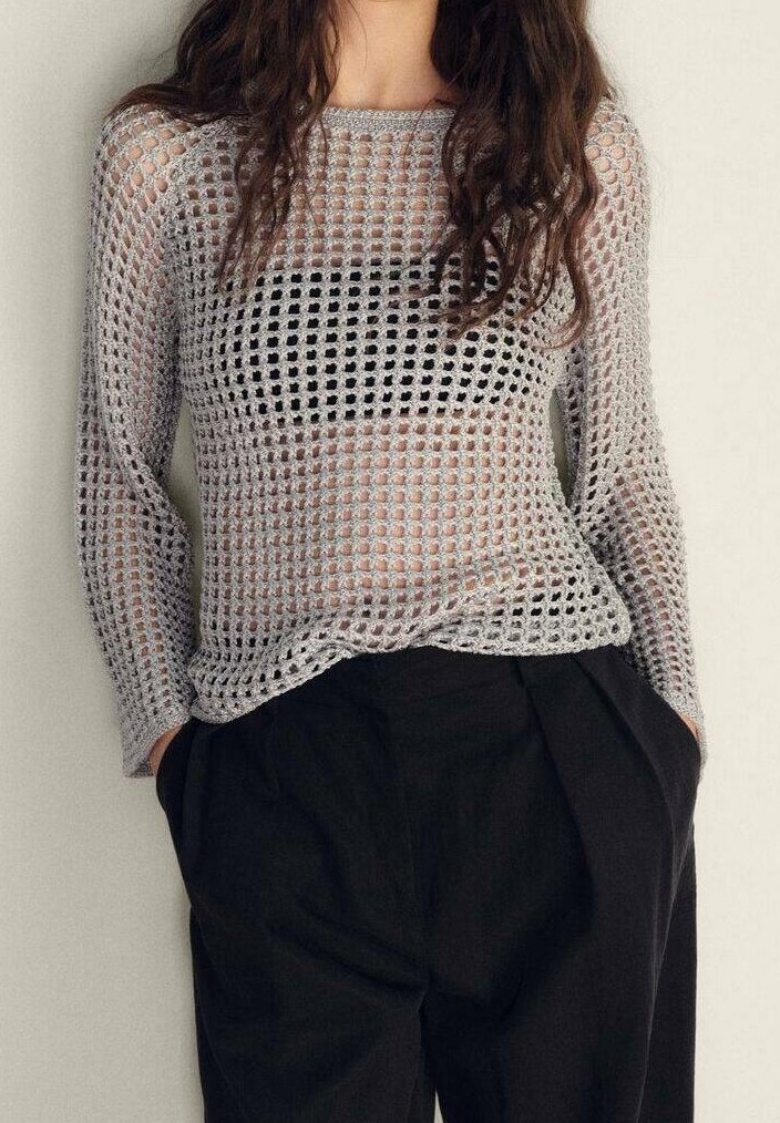 Femme portant un pull gris à manches longues en maille ajourée sur un haut noir, associé à un pantalon noir ample, mains dans les poches.