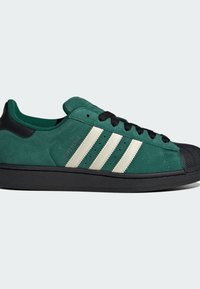 Groene suède sneaker met zwarte accenten, voorzien van een zwarte rubberen neuskap en drie witte strepen. Platte zwarte veters en een gevoerde tong.