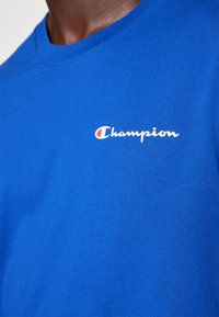 T-shirt en coton bleu avec un col rond et un petit logo "Champion" blanc imprimé sur la poitrine gauche. Coupe lisse et décontractée.