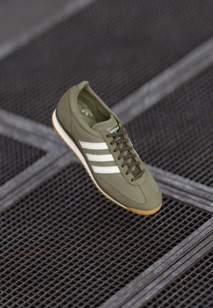 Oliwkowo zielony but sportowy Adidas z białymi paskami i brązową podeszwą, spoczywający na czarnej metalowej powierzchni z siatką, ułożonej diagonalnie.