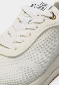 Άσπρο πλεκτό αθλητικό παπούτσι με λευκά κορδόνια, χρυσή καρδιά-οπή και λογότυπο "LOVE MOSCHINO" στη γλώσσα.