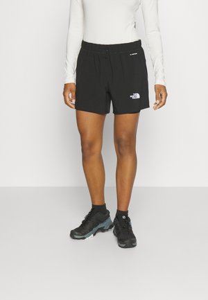 Osoba ubrana w czarne szorty The North Face, czarne buty turystyczne, czarne skarpetki oraz białą bluzkę z długim rękawem stoi na szarej podłodze.