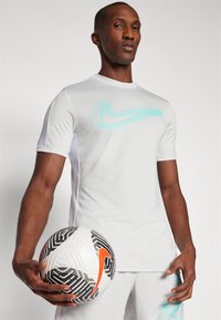 La maglietta sportiva bianca a maniche corte di Nike presenta un logo turchese, un tessuto liscio e dettagli in rete. Un pallone da calcio in bianco e nero è tenuto in basso.