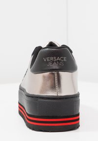 Sneaker Versace Jeans avec un dessus argent métallique, des accents noirs et une semelle noire texturée avec des bandes rouges sur le bas.