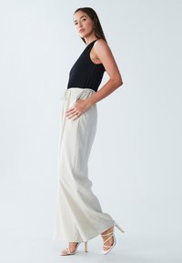 Melnas faktūras tops kopā ar gaiši bēšām plašām biksēm, kurām ir elastīga josta un aukliņa. Modelis nēsā baltus siksnu papēžus.