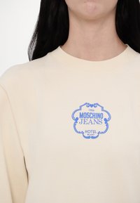 Beige trui met blauw Moschino Jeans Hotel Milano-logo gedrukt op de borst, gedragen door een persoon met lang donker haar.