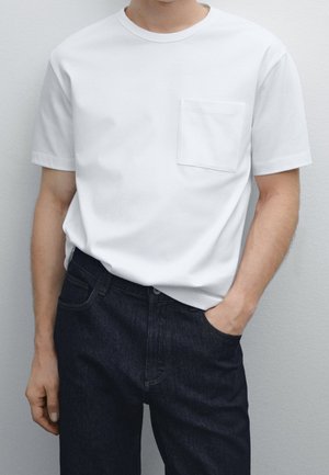 Personne portant un t-shirt blanc uni à manches courtes avec une poche poitrine et un jean bleu foncé, main dans la poche, sur un fond neutre.