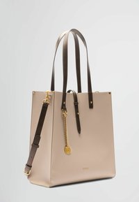 Borsa tote in pelle beige con manici in pelle marrone, decorazioni in oro, fondo piatto e un dettaglio a catena laterale con un pendente per una decorazione aggiuntiva.