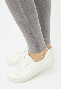 Leggings grises con una textura suave que presentan ocho botones a presión a lo largo de la costura lateral. Combinados con zapatillas blancas sobre un fondo claro.