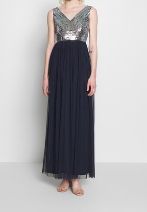 Femme portant une robe de soirée sans manches avec un bustier en V à paillettes argentées et une longue jupe noire fluide, debout devant un fond uni.