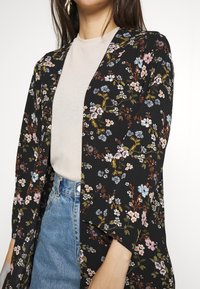 Kvinna som bär en svart blommig långärmad cardigan över en beige topp och blå högmidjade jeans, visad från halsen till höfterna.
