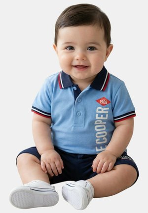 Bébé garçon souriant assis, portant un polo bleu avec le texte "Lee Cooper", un short marine et des baskets blanches sur un fond blanc.