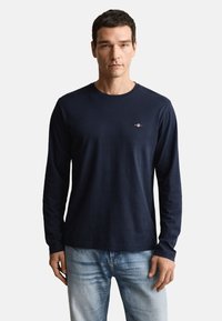 Marineblå langærmet skjorte lavet af bomuld, med rund halsudskæring og et lille logodetalje på brystet. Bæres med lyseblå jeans.