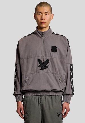 Man met een grijze trui met zwarte arendpatches en ritszakken, gecombineerd met een bijpassende olijfgroene broek, staand tegen een grijze achtergrond.