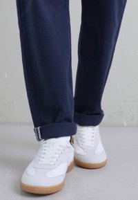 White sneaker with a smooth leather upper, beige rubber sole, and tonal laces. Worn with dark blue tapered pants, showcasing rolled cuffs.