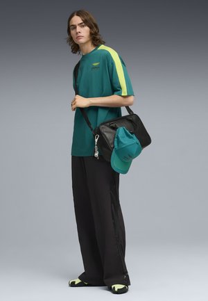 Młoda osoba ubrana w turkusową koszulkę z żółtym paskiem, szerokie czarne spodnie, buty z neonowymi akcentami, nosząca czarną torbę z dopasowaną turkusową czapką przymocowaną.