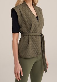 Tilkkutyylinen oliivinvihreä liivi, jossa on kietaisumalli, vyötäröllä vyö ja hihaton tyyli, yhdistettynä oliivinvihreisiin leggingseihin, jotka korostavat sileää tekstuuria.