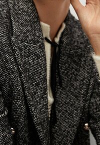 Cappotto tweed grigio e nero con finitura texture, dotato di colletto bianco e pizzo nero, con dettagli a bottoni sulle maniche.