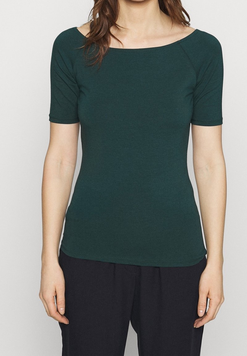 Mörkgrön kortärmad topp med båtkaft, smal passform, mjuk textur och rundad nederkant, bärs med svarta byxor.