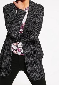 Černo-bílý vzorovaný blazer s uvolněným střih, dlouhými rukávy a otevřeným vpředu, vrstvený přes květinovou košili. Černé kalhoty dotahují vzhled.