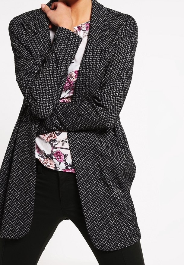 Černo-bílý vzorovaný blazer s uvolněným střih, dlouhými rukávy a otevřeným vpředu, vrstvený přes květinovou košili. Černé kalhoty dotahují vzhled.