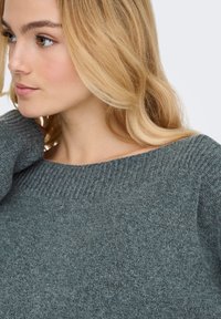 Siv pulover iz pletenine z širokim izrezom in rebrasto teksturo, ki vsebuje mehak material in rahlo padajoče nabore na ramenih.