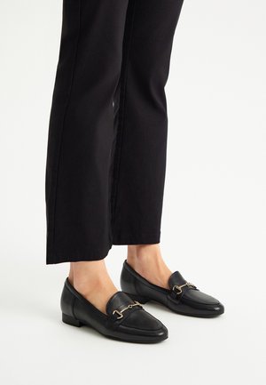 Mocassini neri in pelle con tacco basso, dotati di un accento in fibbia tono oro. Indossati con pantaloni neri a sigaretta, che mettono in risalto un design classico.