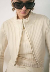 Cream ribbed knit sweater with a high collar and a half-zip closure. Layered with a matching knit top, featuring a textured finish.