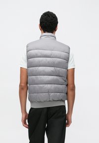 Siva prešita jopič z visokim ovratnikom in vodoravnimi šivi, z rebrastim robom in detajli na ramenih, nošen čez svetlo srajco.