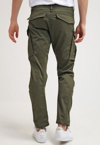 Man som bär olivgröna cargobyxor och vita sneakers, står med ryggen mot kameran mot en enkel bakgrund.