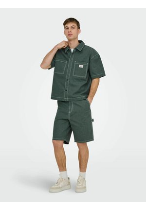Ung mann som har på seg en mørkegrønn kortermet skjorte med knapper og matchende shorts med lyse sømmer, hvite sokker og beige joggesko.