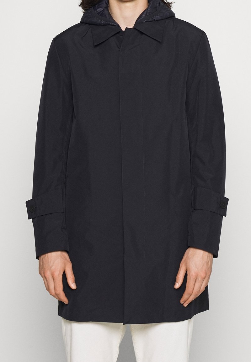 Manteau imperméable bleu marine avec capuche détachable, présentant un design classique, des poignets boutonnés et une texture lisse. Aucun motif ni ornement.