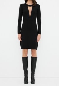 Schwarzes, eng anliegendes langärmeliges Kleid mit tiefem V-Ausschnitt und Ausschnittdetails; kombiniert mit kniefreien schwarzen Lederstiefeln. Glatte Textur.
