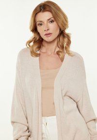 Cardigan lavorato a maglia rosa chiaro con apertura frontale e maniche lunghe, indossato sopra una maglietta beige, abbinato a pantaloni bianchi. Morbido al tatto e vestibilità rilassata.