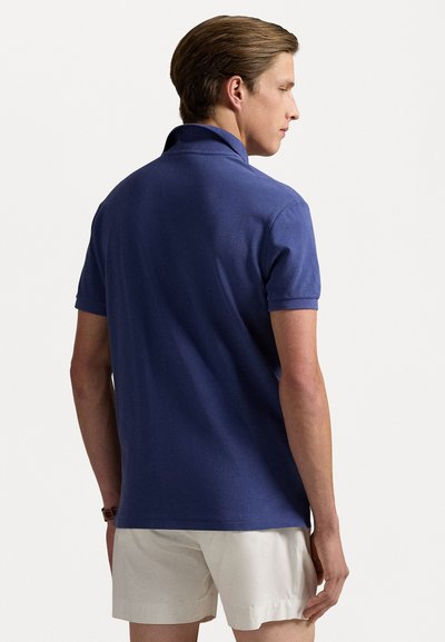 Granatowa koszulka polo z krótkim rękawem, klasycznym kołnierzykiem i ribbowymi mankietami, wykonana z miękkiego, gładkiego materiału. Model nosi jasne szorty.