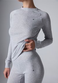 Persona che indossa un top aderente a maniche lunghe grigio chiaro e leggings abbinati, sollevando leggermente il top per rivelare l'elastico in vita.