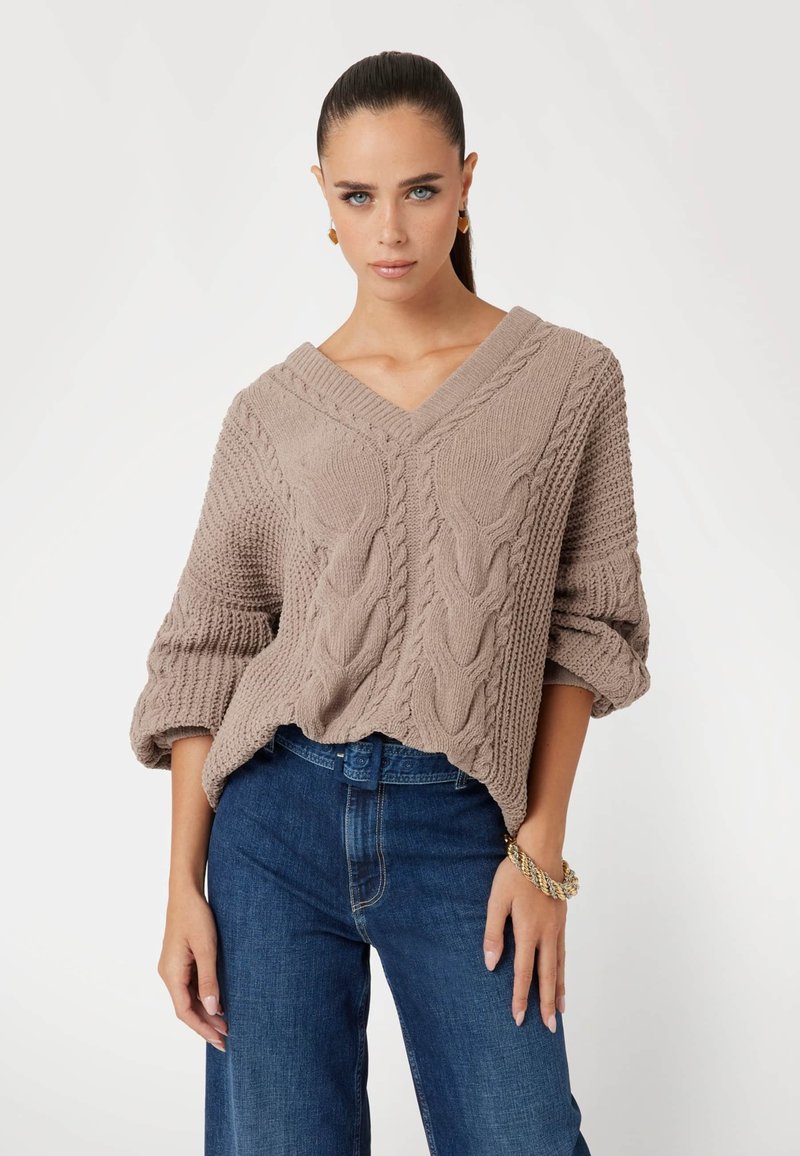 Beige kabeltrui met V-hals, driekwart mouwen en geribbelde textuur, gecombineerd met blauwe denim jeans.