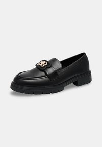 Mocassin en cuir noir avec semelle épaisse et boucle décorative en métal doré sur le dessus, incliné légèrement vers la droite sur un fond blanc.