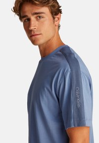 Camiseta de algodón azul para hombre con mangas cortas, cuello redondo, acentos texturizados en los hombros y logo bordado de "Calvin Klein" en la manga.