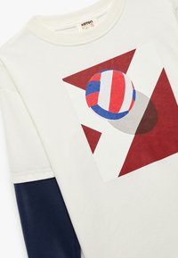 Weißes Baumwoll-T-Shirt mit einem bunten Volleyball-Design auf einem rot-creamfarbenen geometrischen Hintergrund. Marineblaue lange Ärmel.