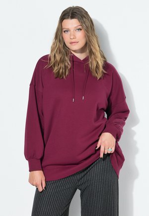 Jeune femme aux cheveux ondulés portant un sweat à capuche bordeaux ample et un pantalon à rayures grises foncées, se tenant devant un fond blanc.