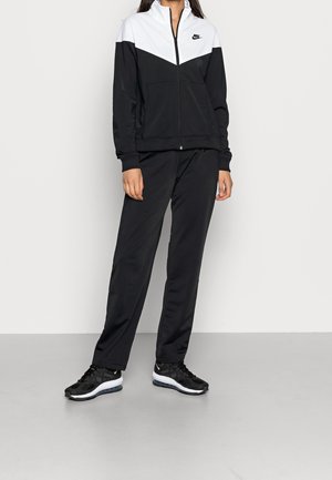 Person wearing a black and white Nike zip-up jacket, black pants, and black Nike sneakers standing against a plain light background.