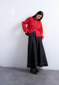 Femme portant un cardigan rouge boutonné, un jean noir à jambes larges et des bottines noires, debout contre un mur blanc uni.